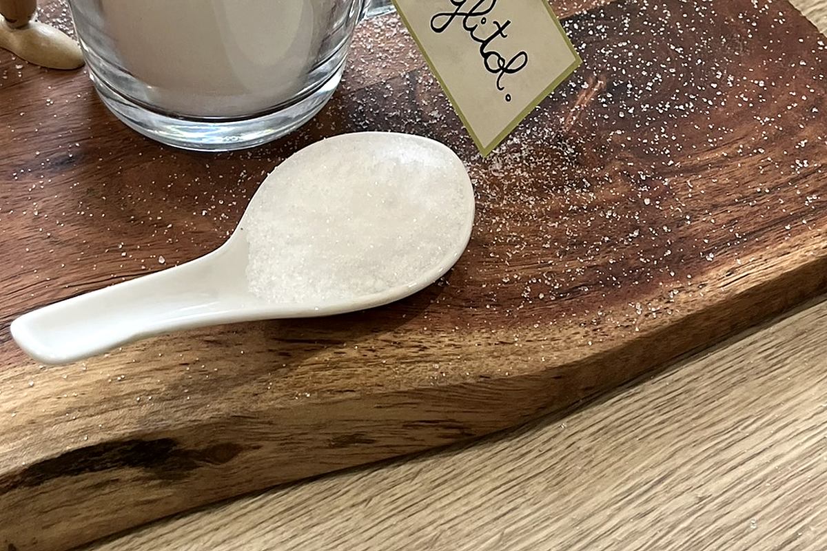 Une cuillère bombée de xylitol posée sur une planche en bois, à côté d'une tasse en verre remplie de cristaux