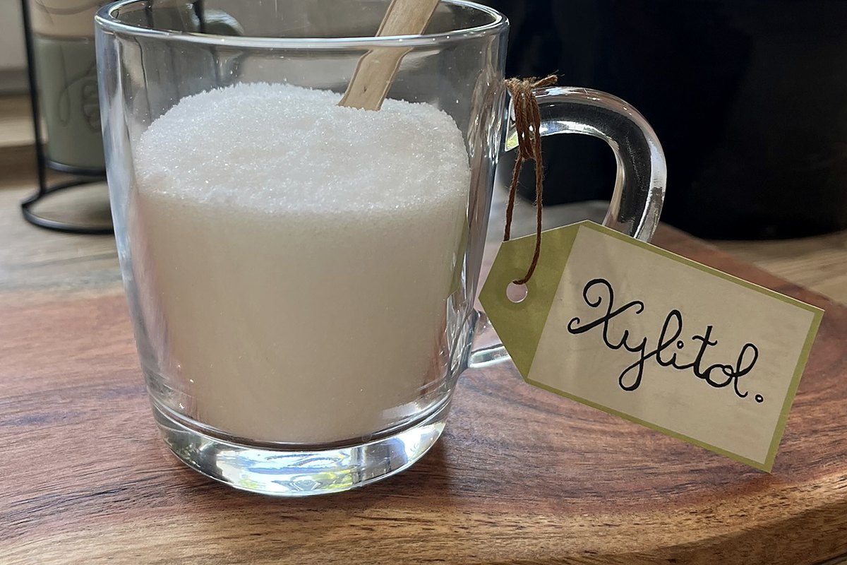 Tasse en verre remplie de xylitol, une étiquette manuscrite attachée à l'anse, devant une fenêtre